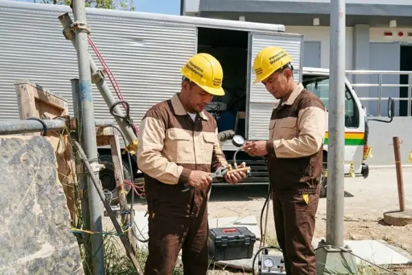 Two plumbing technicians in tan and brown uniforms and yellow hard hats working together outdoors. One technician uses a wrench on a brass valve assembly while the other monitors a pressure gauge. A service truck with an open rear compartment is parked behind them. A worker of Visayas Malabanan Negros Occidental, Bacolod City, Sum-ag.