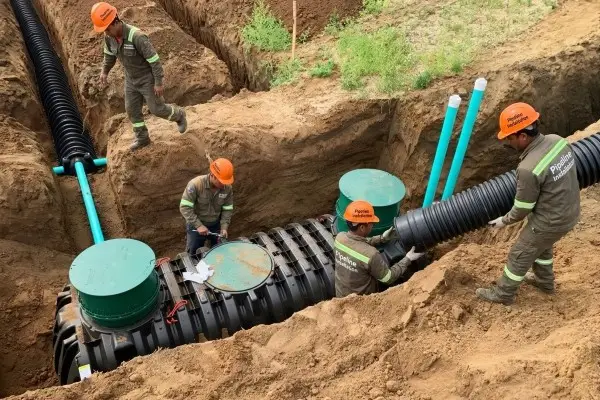 A team of four technicians in grey workwear and orange hard hats installing a large underground septic or STP tank system in Dumaguete. The crew is shown connecting heavy-duty corrugated black piping and teal access risers within a deep excavated trench.
