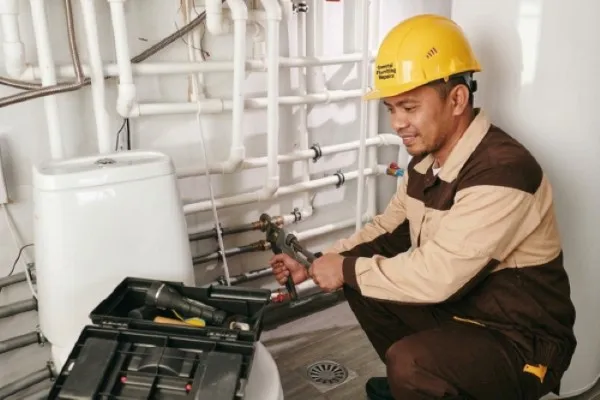 A smiling plumber in a brown and tan uniform and yellow hard hat uses a large pipe wrench to work on an intricate wall-mounted piping system. An open black toolbox sits in the foreground as the technician performs a residential or commercial plumbing installation near a white toilet tank. A worker of Visayas Malabanan Negros Occidental, Bacolod City, Sum-ag