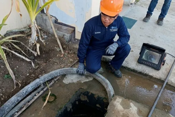 A professional plumber in a dark blue uniform and orange hard hat performs a CCTV pipe inspection. He is kneeling next to an open drain while monitoring a portable digital screen that displays a real-time view from inside the plumbing line. A worker of Visayas Malabanan Negros Occidental, Bacolod City, Sum-ag